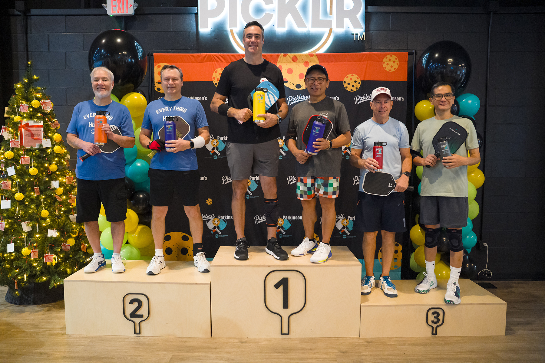 Five men stand on a winners podium (1st, 2nd, 3rd) holding water bottles and paddles at a pickleball event, with a festive backdrop and Christmas tree nearby.