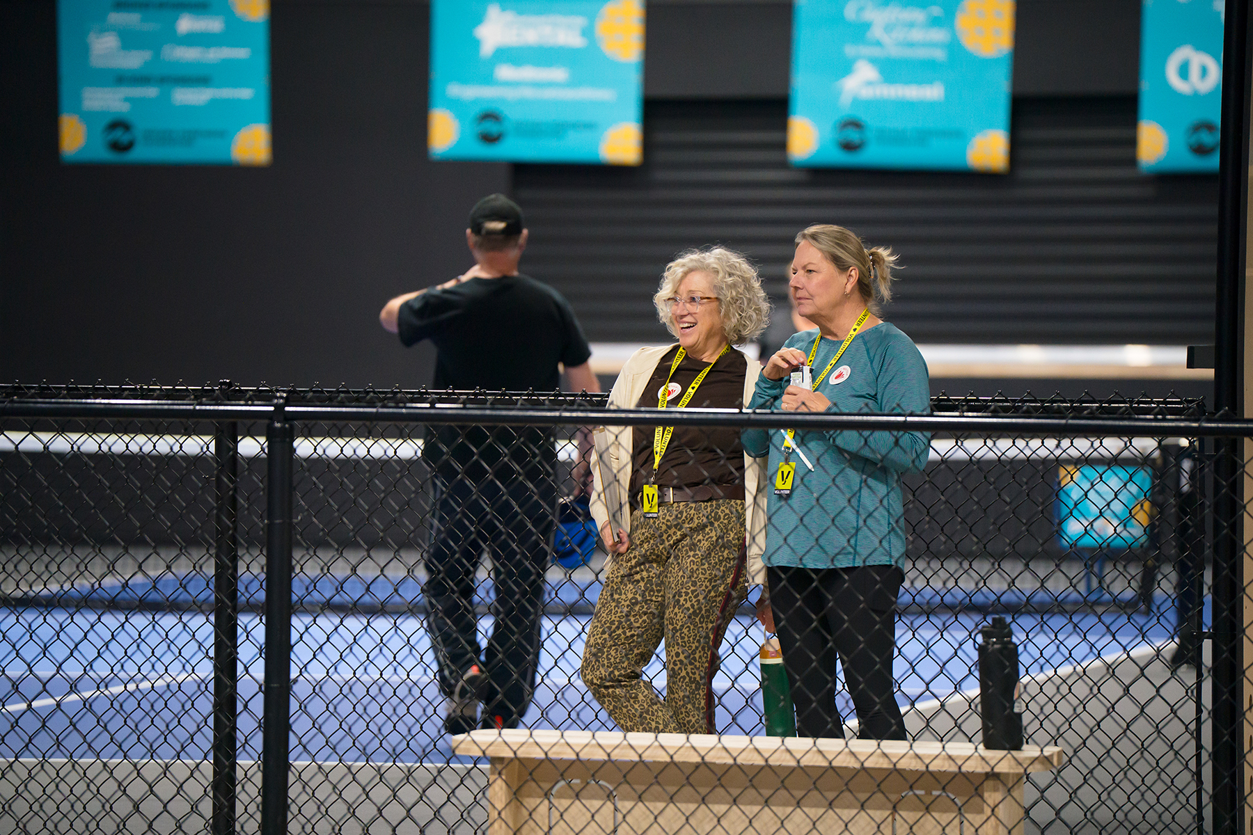 Two women with yellow lanyards standing behind a chain-link fence in an indoor batting cage, chatting as blue mats and banners hang in the background,