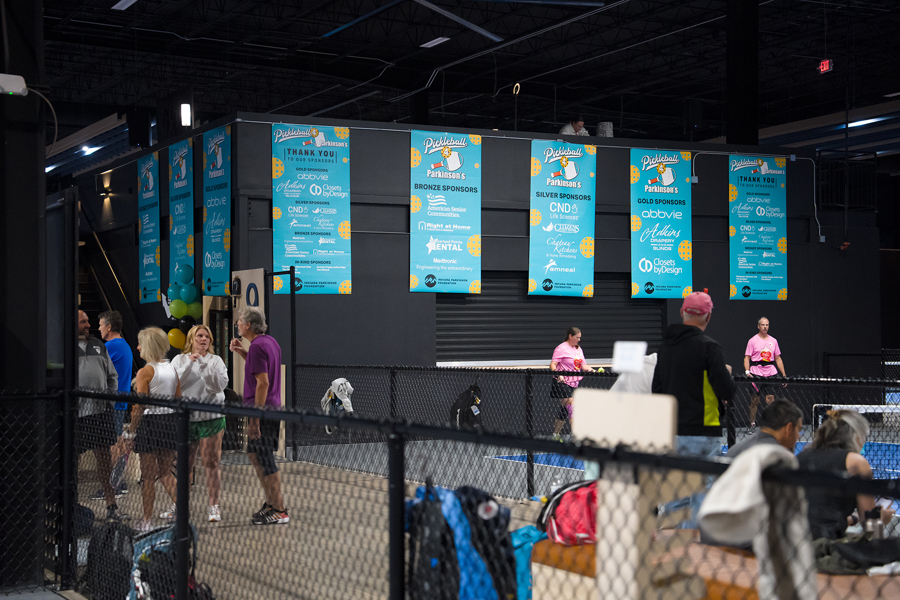 Indoor sports venue with blue sponsor banners reading Pickleball and Parkinson's hanging above a fenced pickleball court; people stand nearby, some in pink shirts preparing to play.