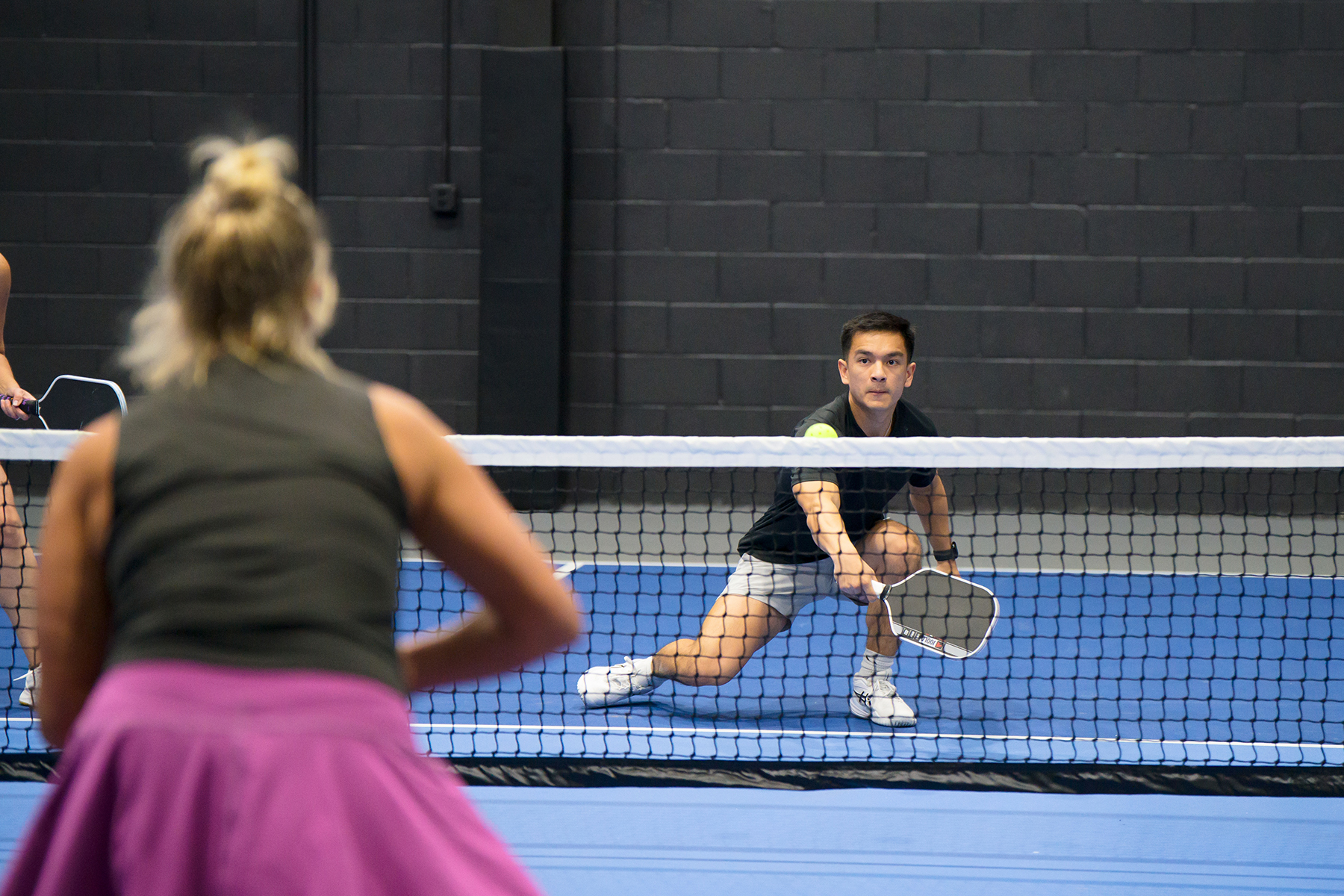 Two people play indoor tennis across a net; the foreground player, wearing a pink skirt, faces away from the camera as the other player lunges to return the ball near the net.