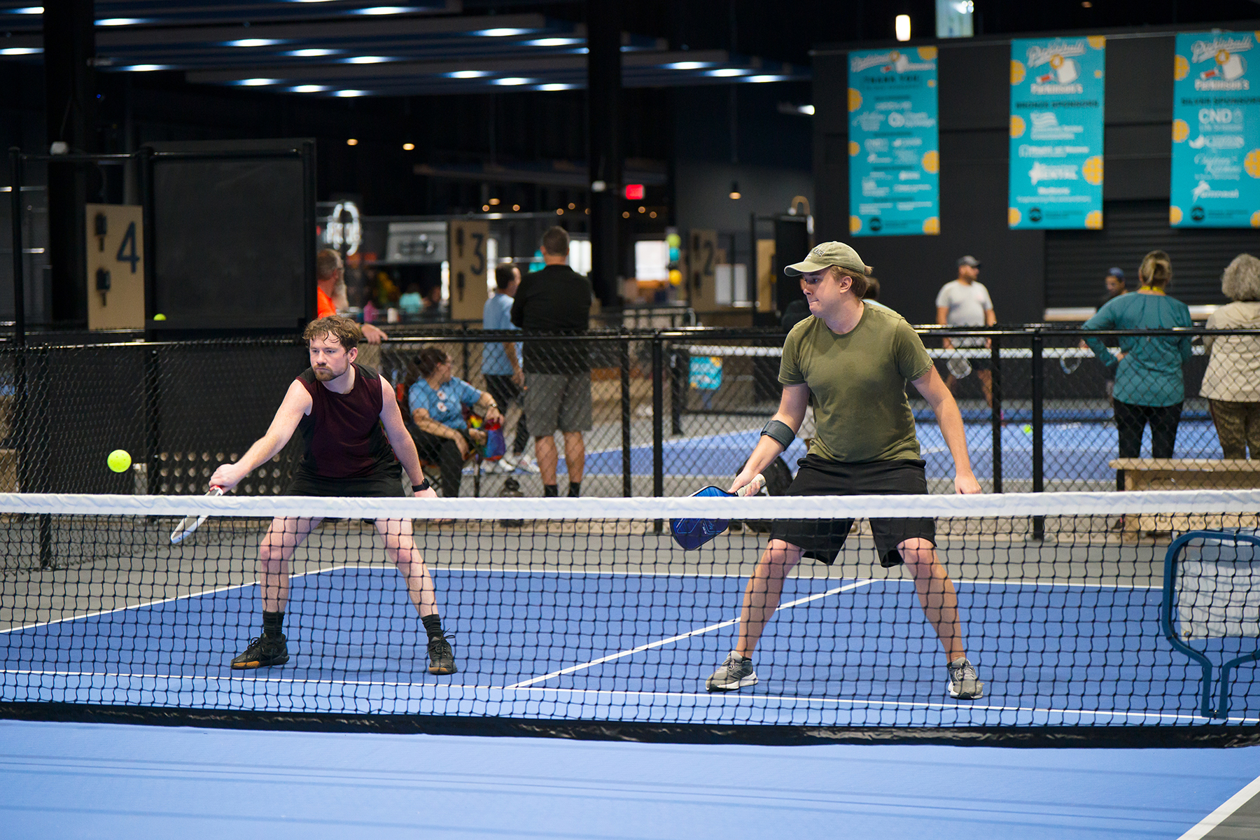 Two men play pickleball on a blue indoor court; one in a maroon sleeveless shirt reaches for the ball at the net as the other prepares a return with a paddle nearby.